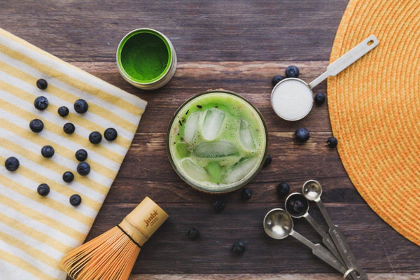 Blueberries, a Junbi Matcha whisk, measuring spoons and a matcha tin are sitting on a table surrounding a blueberry matcha latte.