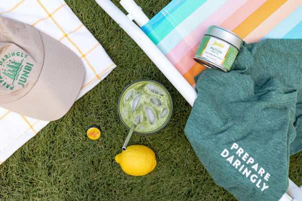 Junbi hat, matcha tin and a green shirt that says 'Prepare Daringly" laying on the grass. With a matcha tea and a lemon next to it, representing "How to Make Sparkling Matcha Lemonade at Home"